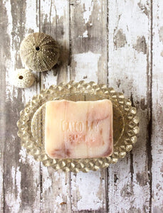 a creamy white bar of soap with borwn and pink swirls sits in a gold soap dish