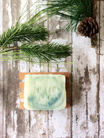 an off white bar of soap with green swirls sits in a wood soap dish surrounded by pine boughs