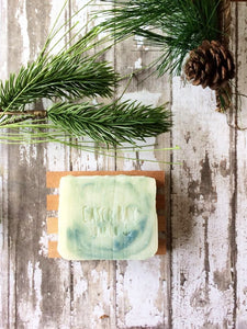 off white bar of soap with green swirls sits in a wood soap dish suronded with pine boughs