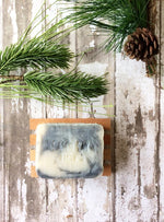 a creamy white bar of soap with grey swirls sits in a wooden soap dish surounded by pine boughs
