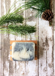 a creamy white bar of soap with grey swirls sits in a wood soap dish surrounded by pine cones and boughs