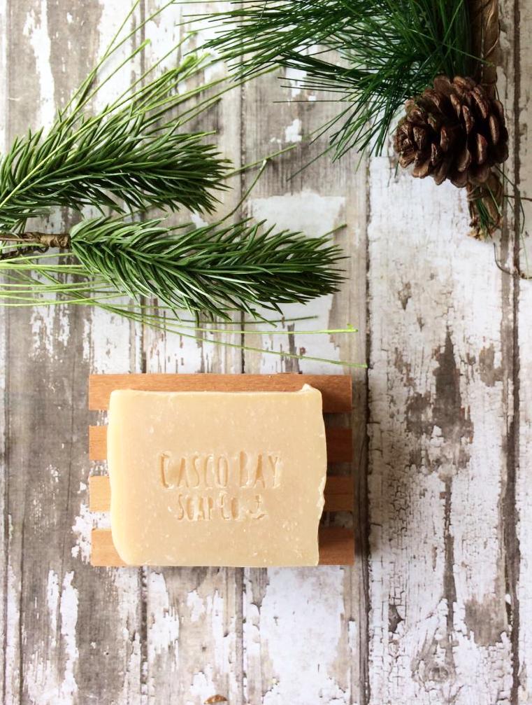 a tan bar of soap sits in a wood soap dish surrounded by pine boughs