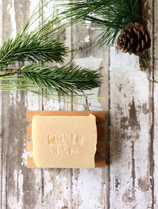 a tan bar of soap sits in a wood soap dish surrounded by pine boughs