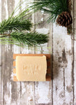 a yellowish tan bar of soap sits in a wood soap dish surounded by pine boughs