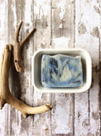 creamy white bar of soap with blue and black swirls sits in a white soap dish