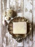 a creamy white bar of soap with light purple swirls sits in a silver shell shaped soap dish