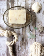 a creamy white bar of soap with oatmeal flakes in it sits in a glass soap dish