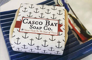 a bar of soap with a paper soap label that has blue anchors on it sits in a blue glass soap dish
